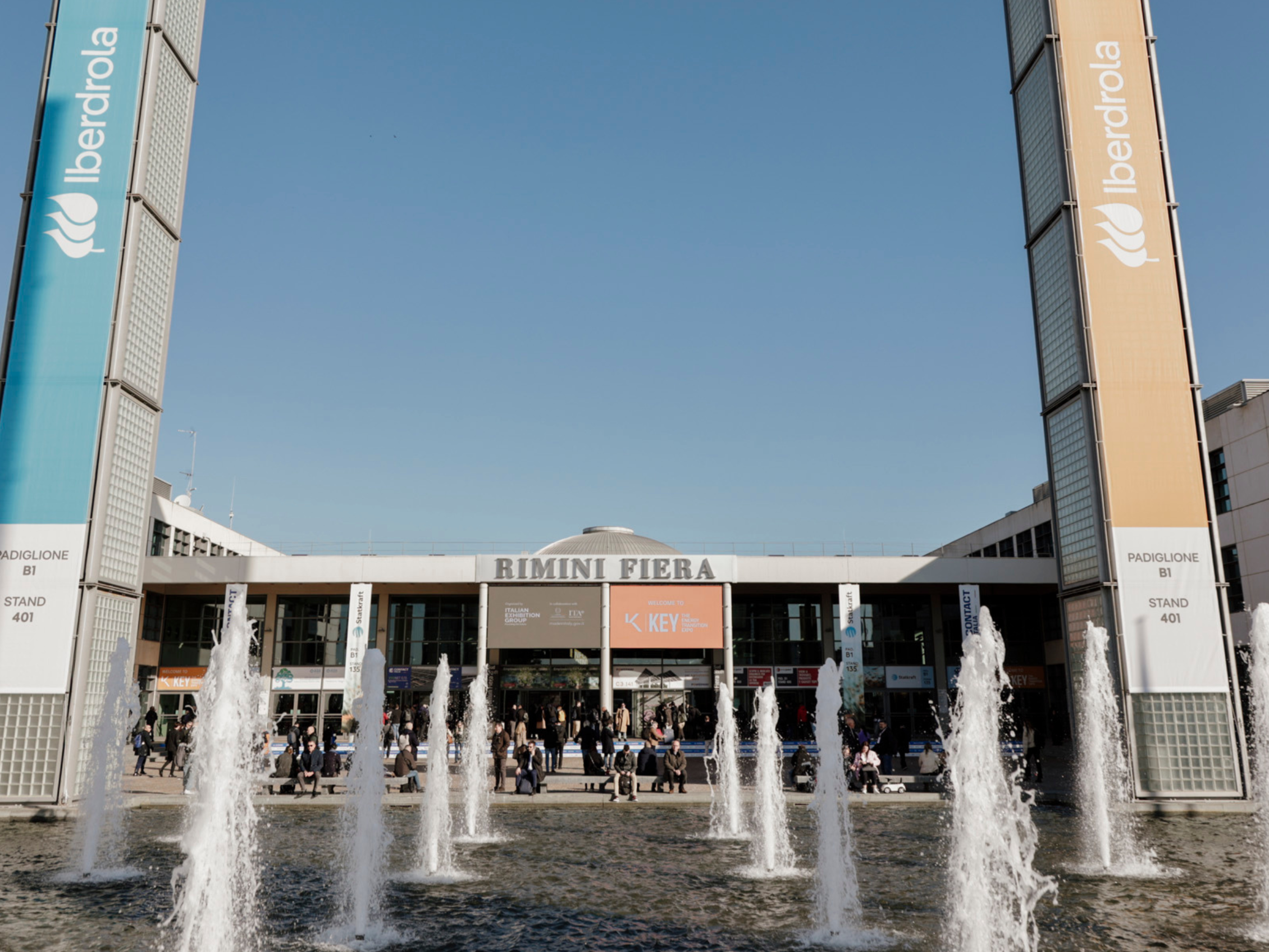 south entrance of the Rimini Expo Centre with many visitors outside, a pool with fountains, and large screens displaying the K.E.Y. logo and useful information