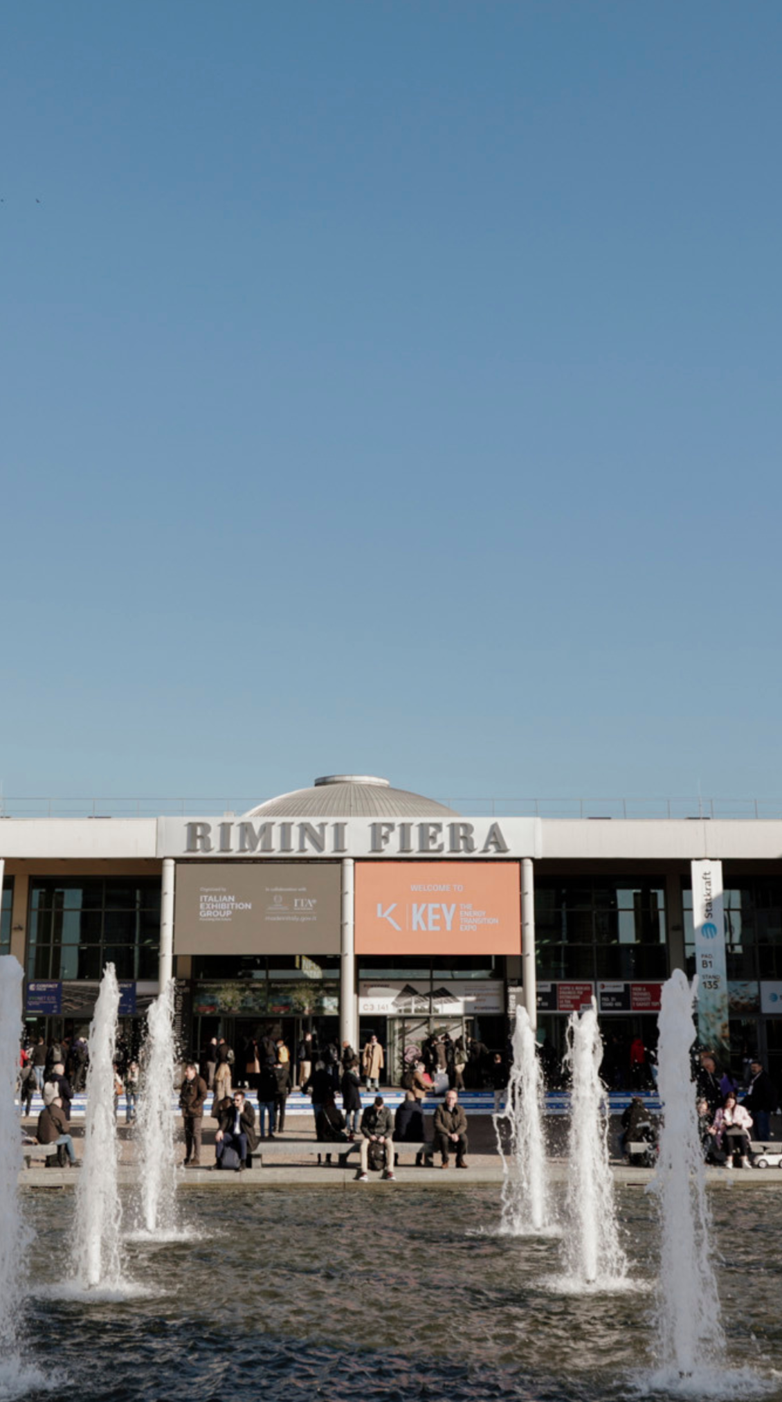 south entrance of the Rimini Expo Centre with many visitors outside, a pool with fountains, and large screens displaying the K.E.Y. logo and useful information