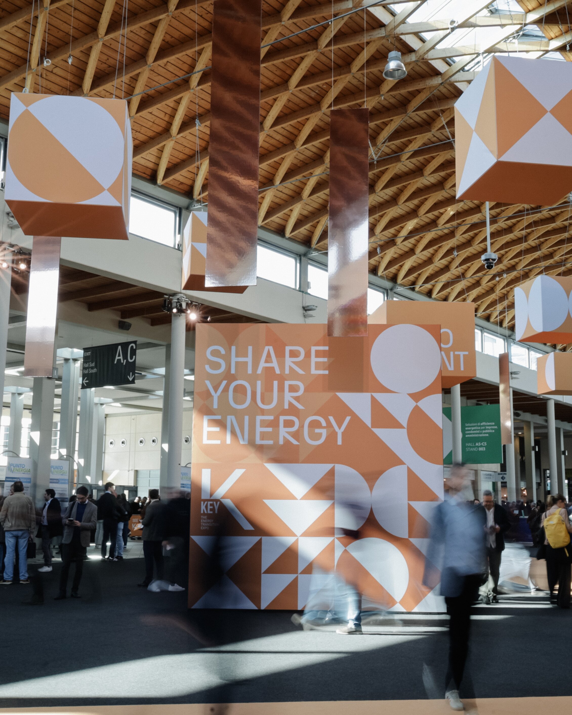South Hall of the Rimini Expo Centre during K.E.Y. featuring installations with hanging cubes decorated with orange geometric graphics. At the center, a large cube displays the text 'SHARE YOUR ENERGY'