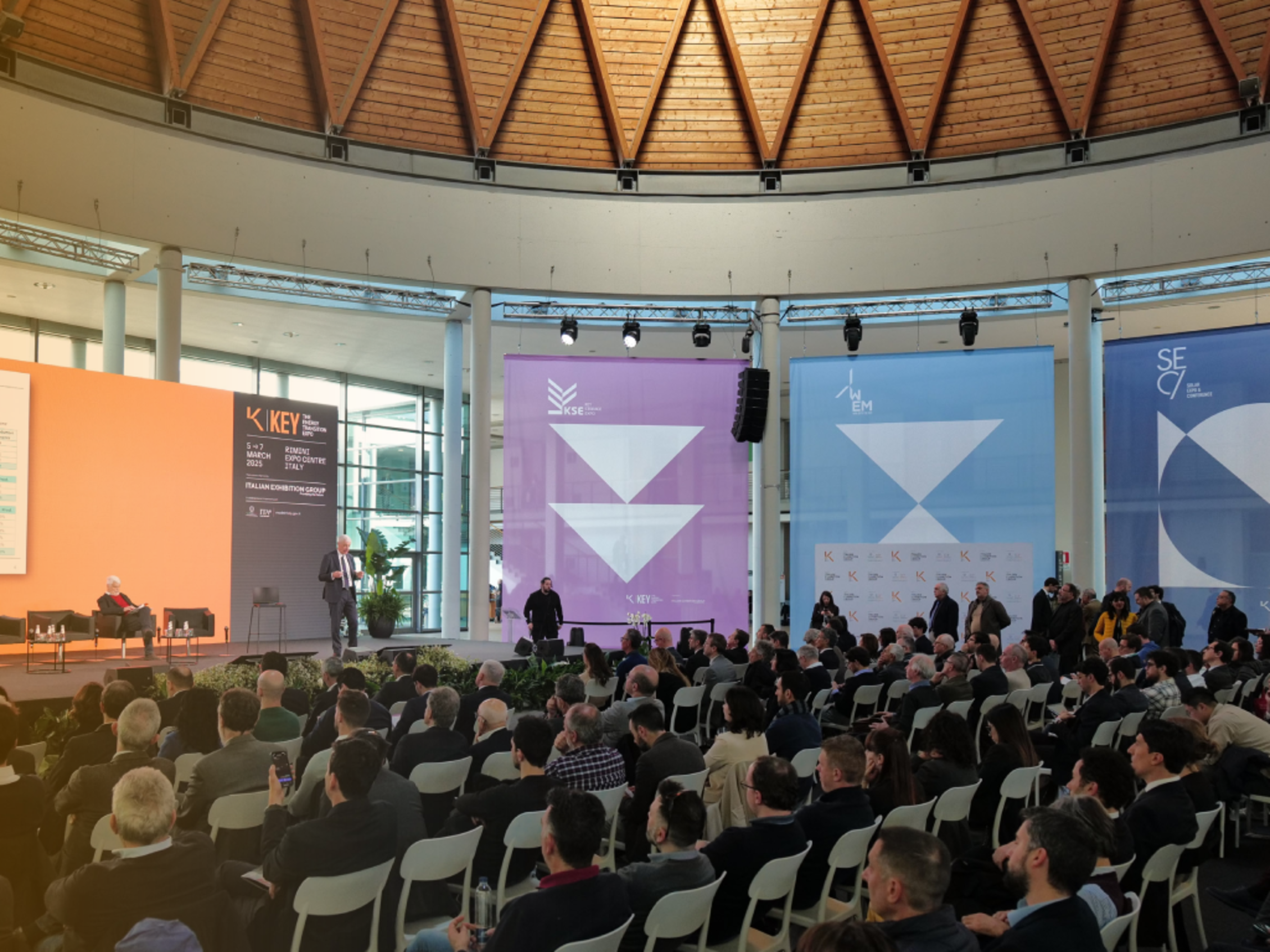 Conference in the Lorenzo Cagnoni Dome at the south entrance of the Rimini Expo Centre during The Energy Transition Expo, with many seated attendees, a speaker on stage talking in front of an orange screen, and large colorful panels in the background