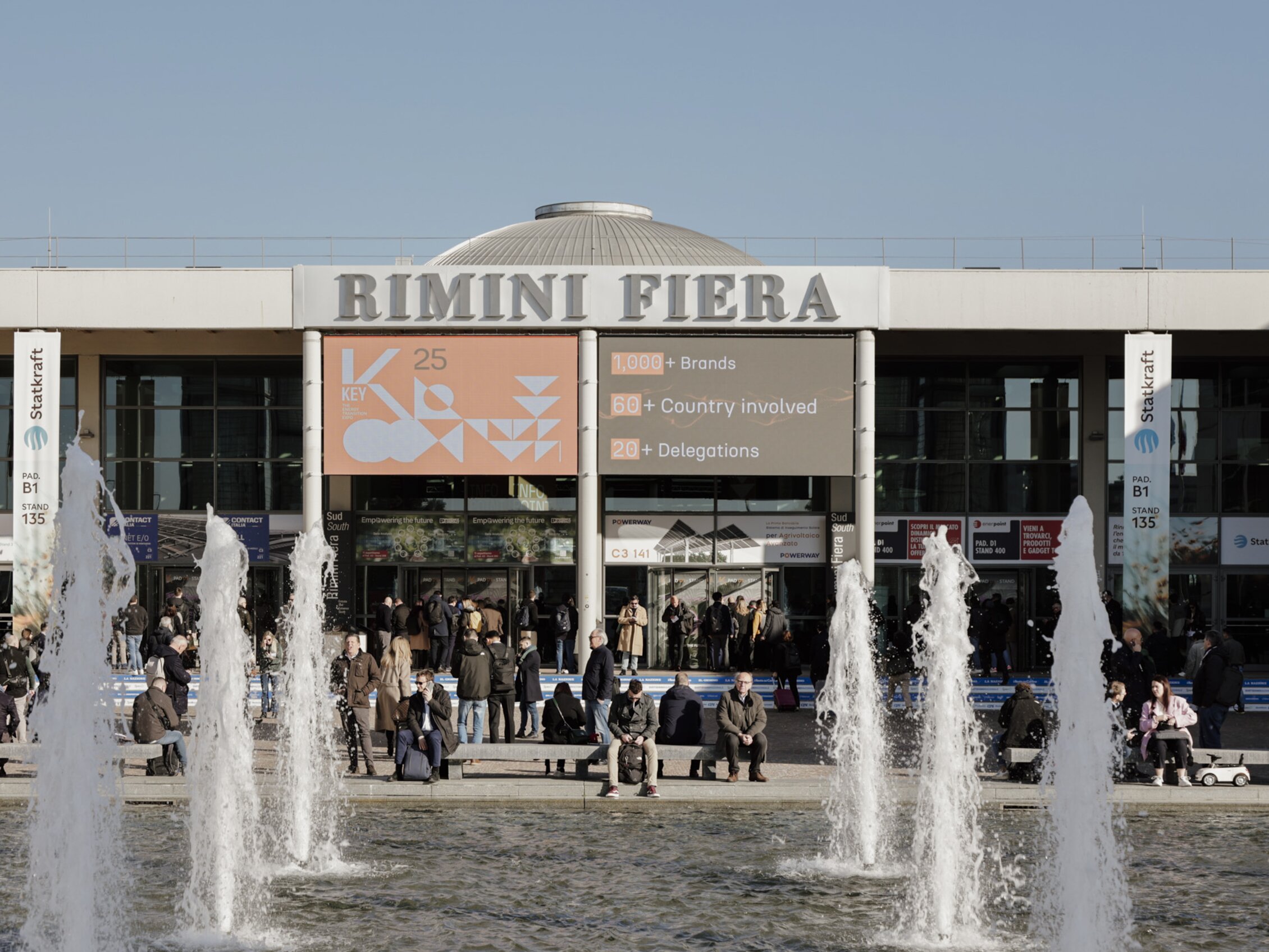 Main entrance of the Rimini Expo Centre during K.EY - The Energy Transition Expo, with many visitors outside, a pool with fountains, and large screens displaying the K.EY logo and useful information