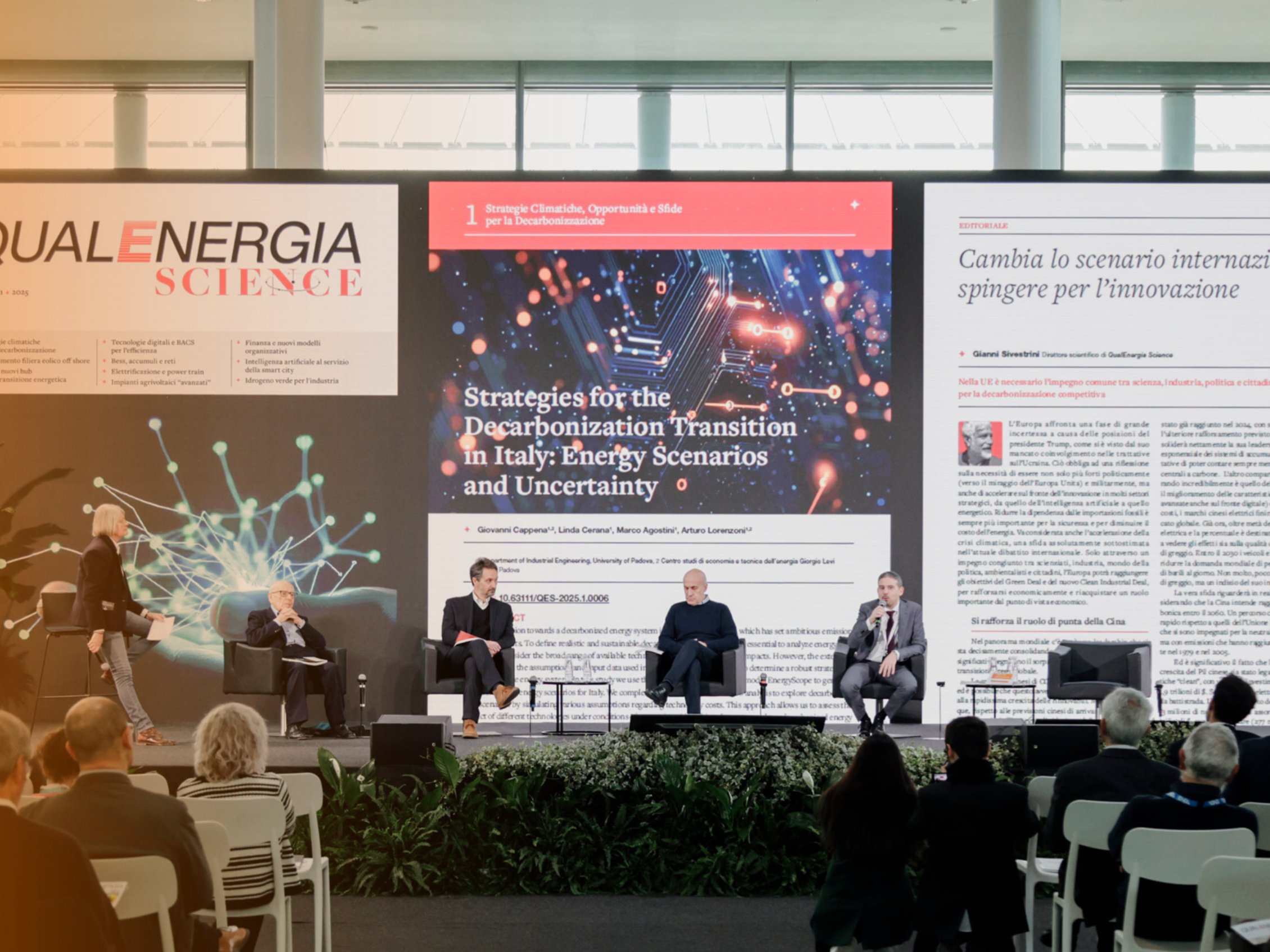 Panel of experts on stage at a conference about strategies for the decarbonization transition in Italy. In the background, three large screens display the logo of the magazine 'QUALENERGIA SCIENCE' and an article titled 'Changing the international scenario, pushing for innovation'
