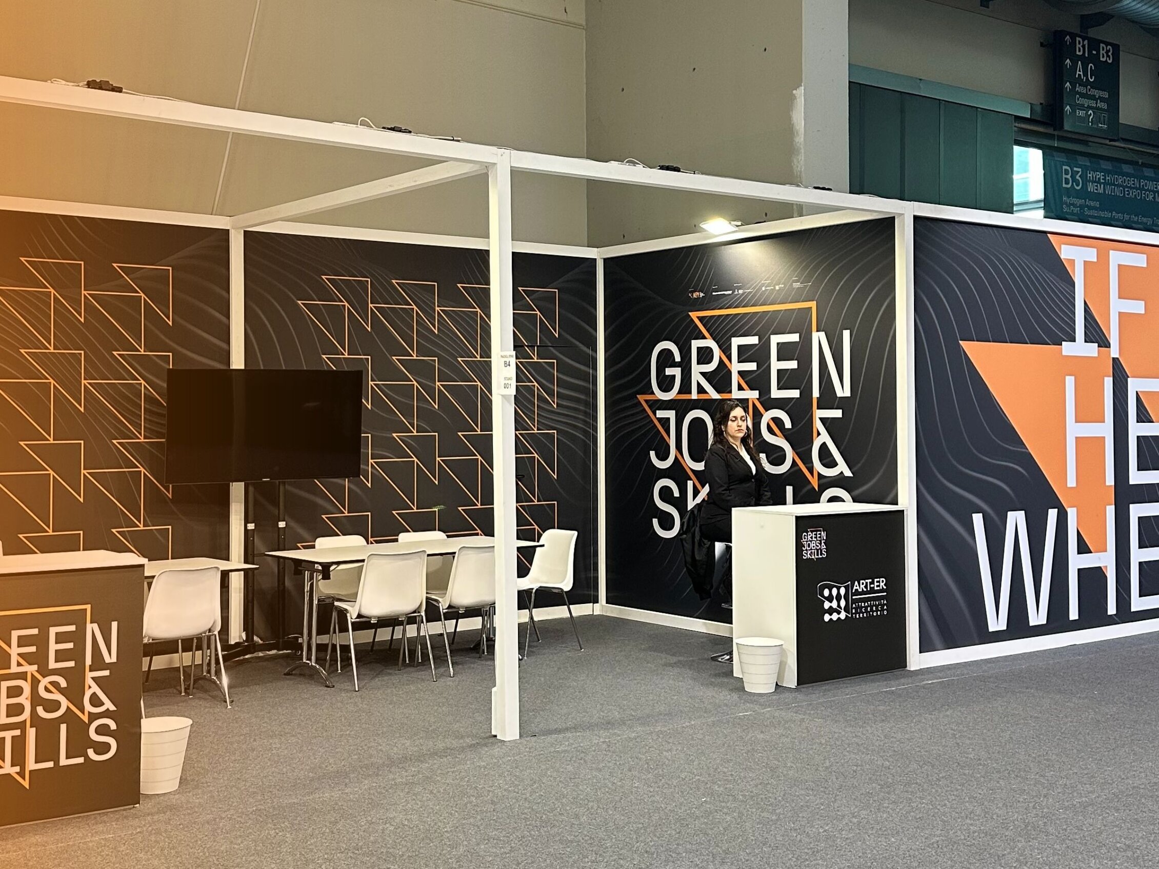 Green, jobs and skills booth with tables surrounded by chairs, a host and a large slogan on the wall that reads 'if not here where?'